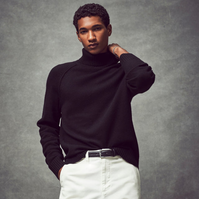 A photo of a man looking at the camera in a studio, wearing a dark cashmere turtleneck and white pants.