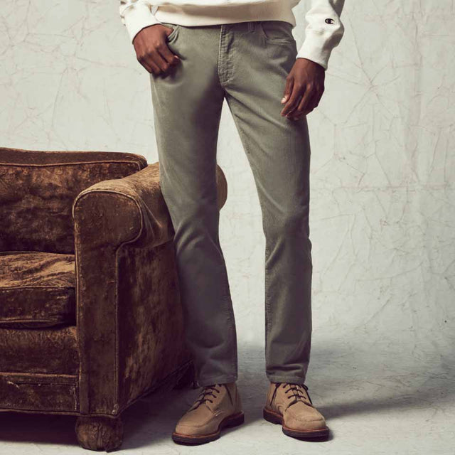 A close-up photo of a man in pair of corduroy 5-pocket chinos and tan boots, standing in a studio next to a couch, with one hand in his pocket.