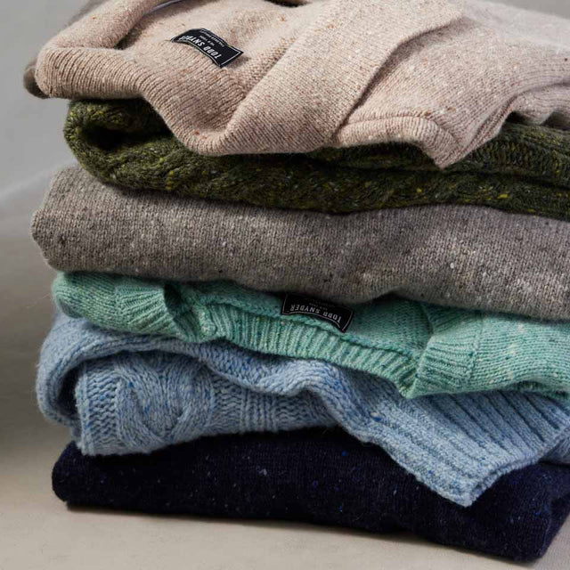 A close-up photograph of a stack of wool sweaters in muted colors, all knit from Donegal yarn.